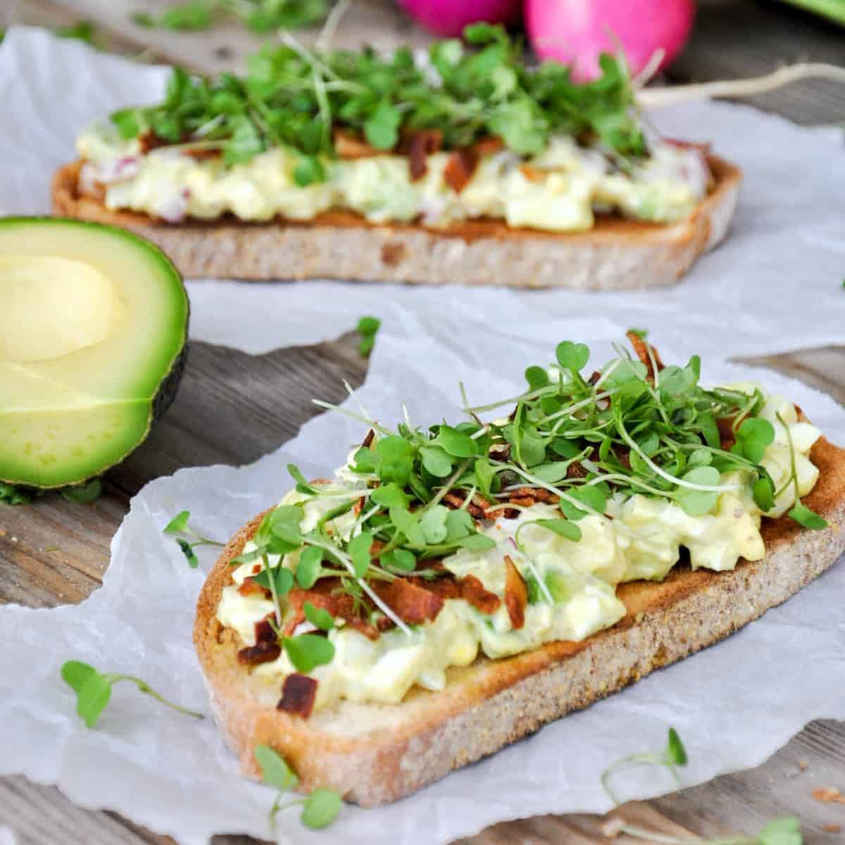 Two slices of toasted sourdough topped with avocado and egg, bacon bits, and microgreens